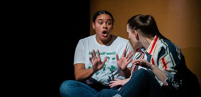 2 actors sitting on stage looking at each other and talking with one holding her hands up and her mouth open.